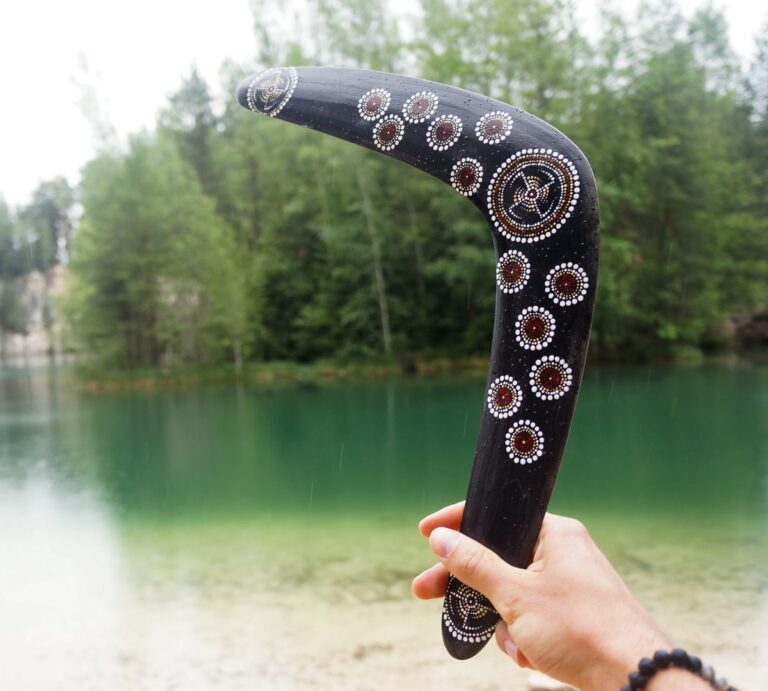 En træ boomerang bliver holdt ud mod en sø. Boomerangen er flot udsmykket med blomster. Billedet er meget drømmende.