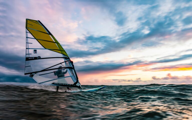 mads schmidt rasmussen 1aFbxEn2rqI unsplash Person er ude og windsurfe i de stille bølger og med en smuk aften himmel med røde, violette og orange farver.